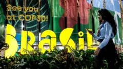 Mulher passando por letreiro que diz: Vejo você na COP 30 Brasil