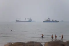 Buques de carga y petroleros permanecen en primer plano frente a la costa, mientras que personas nadan y se refrescan en las aguas costeras en un día brumoso.