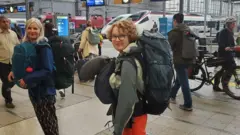 Una adolescente en una estación de tren con una mochila grande a la espalda y otra más pequeña al frente.