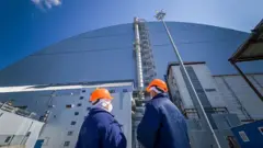 Чорнобильська катастрофа арка укриття перед якою у помаранчевих шоломах стоять двоє працівників 