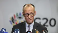 Merz takım elbiseli ve mikrofonların olduğu kürsüde ayakta konuşuyor. 22 Kasım 2025. 