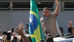 Bolsonaro sorrindo acena para apoiadores diantepin up casino bônusbandeira do Brasil