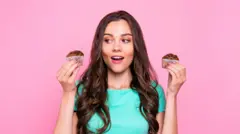 Turkuaz renkli tişört giymiş uzun saçlı bir genç kadın iki elinde tuttuğu muffinleri yukarı kaldırmış bakıyor