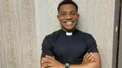 Pasitọ Jerry Eze wọ asọ Alufa dudu, o so kola mọ ọrun, o ka ọwọ mejeeji bo aya, o si n rẹrin