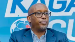 Umuyobozi wa FERWAFA Shema Fabrice yambaye ikositimu y'ubururu n'indorerwamo areba imbere ahagana hejuru. Imbere y'ikoti yambayemo ishati y'umweru n'ubururu. Inyuma ye hari icyapa cy'ubururu cyanditseho mu mweru n'ubururu. 