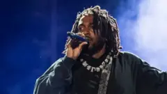 Rapper Kendrick Lamar, wearing black clothes, a necklace, and a bag over his shoulder, holds a microphone as he performs onstage during day three of Rolling Loud Miami 2022 at Hard Rock Stadium on July 24, 2022 in Miami Gardens, Florida