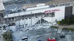 Vista aérea de um shopping center da rede Aeon com parte da fachada destruída, janelas estilhaçadas e destroços espalhados pelo estacionamento e pelas vias ao redor. Alguns veículos e equipes de emergência aparecem na área.