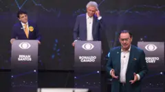 Em um palco de debate, três candidatos aparecem diante de púlpitos identificados com seus nomes. À esquerda, Renan Santos observa a cena; ao centro, Ronaldo Caiado leva a mão ao ouvido; e, em primeiro plano, Augusto Cury fala gesticulando. O fundo exibe gráficos em tons azulados e elementos visuais do evento.