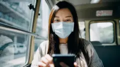 Maskeli ve cep telefonuna bakan bir kadın 