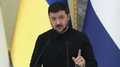 Володимир Зеленський у чорному костюмі стоїть за стійкою на фоні прапорів 