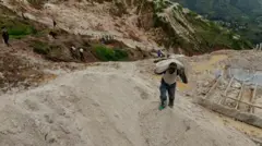 Un mineur transporte un sac de minerai à la mine de Rubaya. Le terrain est vallonné. Derrière lui, on aperçoit d'autres mineurs et, au loin, des champs verdoyants.