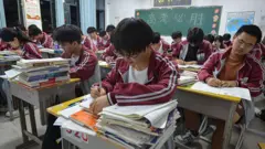 Estudantes do ensino médio na China sentados em suas carteiras, escrevendo, com pilhas de livros ao lado.