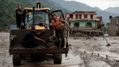 Rescuers use one digger truck to carry an elderly woman to safety. Buildings swamped by mud can be seen in the background
