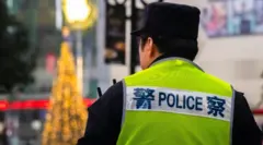  A police officer patrols near an illuminated Apple logo and a festive Christmas tree in the Jiefangbei commercial district on December 17, 2024 in Chongqing, China. He is wearing a bright yellow vest with police written on it in both Mandarin and English. Christmas tree lights can be seen in the background