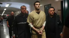 Luigi Mangione caminhando, com uniforme de presidiário e algemas, escoltado por dois guardas.