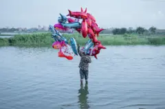 Un hombre de pie en un río, sosteniendo globos de colores vivos por encima de su cabeza.