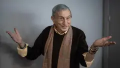 Haldun Dormen, siyah kazak ve kahverengi, desenli bir atkı giymiş halde ellerini iki yana açıp, gülümserken 