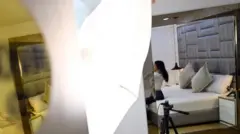 Una chica se prepara para hacer una sesión por webcam en una habitación: se le ve sentada en la cama de espaldas, mientras se arregla el cuello de la camisa antes de salir en vivo. El rostro está desenfocado.