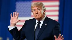 A imagem mostra Donald Trump, um homem de terno escuro e gravata, diante de um fundo desfocado com a bandeira dos Estados Unidos. Ele está com as duas mãos erguidas, especialmente a esquerda aberta em direção à câmera. Sua expressão é séria e contida, e a composição destaca a figura em primeiro plano, com iluminação uniforme e foco nítido no rosto e nas mãos.