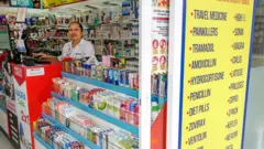 Seorang perempuan berbaju putih sedang berada di balik etalase obat-obatan.