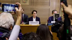 Cámaras de celular toman imágenes de la directora del Louvre sentada ante un escritorio y acompañada de dos hombres