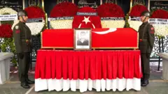 Er Hayrullah Halit Karaman'ın tabutunun iki yanında asker duruyor. Arkada çelenkler görülüyor.