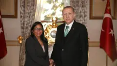 Rodriguez ve Erdoğan arkada Türk bayrağ görünen bir odada el sıkışarak kameraya bakıyor. 