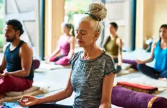 Imagem de arquivo mostra uma pessoa sentada no chão de um estúdio de ioga, praticando meditação, com outras pessoas ao fundo