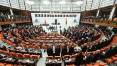 TBMM Genel Kurulu'ndaki milletvekilleri ayakta bekliyor. Arkalarında turuncu sandalyeler ve kahverengi masalar görünüyor. Kürsünün arkasında beyaz arka plan üzerine 'Egemenlik Kayıtsız Şartsız Milletindir" yazıyor.
