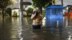 Uma mulher durante a noite em uma rua inundada, com a água quase na altura da cintura e usando um casaco por causa do inverno, em um subúrbio de Santiago, no Chile, durante as enchentes de julho de 2026.