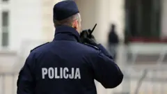 Працівник польської поліції розмовляє по рації. На спині форменого одягу видно напис «POLICJA».