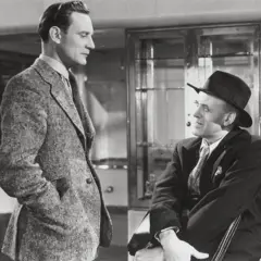 Dos hombres se miran a los ojos. Uno está sentado mientras el otro está de pie frente a él. Ambos llevan traje y corbata. El que está sentado tiene un sombrero. La imagen es en blanco y negro.