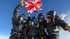 Cuatro escaladores juntos, vestidos con equipo de escalada y máscaras, parados en una cima mientras uno sostiene una bandera del Reino Unido