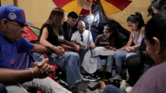 Varias personas sentadas en un campamento improvisado bajo una sombrilla.