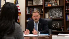 Foto de Brandon Guffey sentado atrás de uma mesa, com estantes de livros e uma bandeira dos EUA ao fundo. Ele tem cabelo curto e claro, barba, e veste terno azul-marinho com gravata azul-clara. À esquerda da imagem está Tir, de costas, com cabelo longo e escuro e um suéter cinza