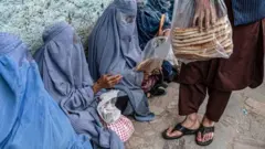 تصویر شماری از زنان افغان که از دست یک مرد نان خشک می‌گیرند. این زنان چادری ابی به تن دارند و در یک صف برای دریافت نان نشسته اند. 