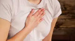 A woman is holding her hand on her chest to indicate a problem with her heart. She is wearing a light pink T-shirt