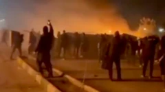 Uma captura de vídeo borrada mostra manifestantes e fumaça de um incêndio; uma pessoa aparece erguendo um celular para registrar os acontecimentos. A imagem foi feita em Mashhad, no início de janeiro