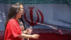 Tahran'da arkada İran bayrağı görünen bir sokakta iki kadın yürüyor. Kadınların başı açık ve biri kırmızı tişört giyiyor.