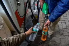 Um frentista vestindo uma jaqueta puffer azul enche uma garrafa plástica com gasolina em um posto de combustível, enquanto a outra mão segura uma garrafa vazia.