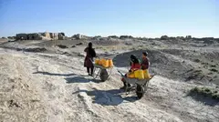 تصویر دو کودک و یک زن که با استفاده از کراچی و بشکه‌ها آب را به خانه‌های شان انتقال می‌دهند. افغانستان در سال گذشته شاهد خشک‌سالی و کمبود آب بود. 