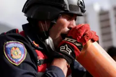 Un rescatista con casco, uniforme, guantes rojos y un tapabocas mal puesto, que lleva debajo de la barbilla, grita por un tubo color naranja de PVC.