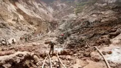 A man works at an open shaft at the mine near Rubaya in 2019. The terrain is hilly and other miners can be seen at a distance.
