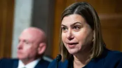 青いスーツを着た、ブロンドをボブカットにした女性が、マイクに向かって話している。背後にはスーツを着た男性の姿がぼやけて写っている