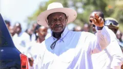 Пожилой мужчина с усами, в светлой шляпе и широкой белой рубашке, с приветственно поднятой рукой