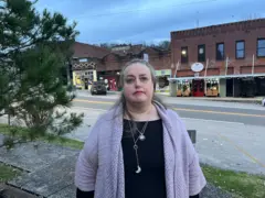 Una mujer con el pelo gris y una rebeca morada está de pie frente a una hilera de tiendas en la plaza principal de Gainesboro.