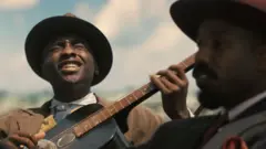 Imagem do filme 'Pecadores' mostrando Miles Caton como Sammie tocando violão e Michael B. Jordan olhando para frente