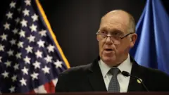 アメリカ国旗と米国土安全保障省の旗を背に、演台のマイクに向かうスーツ姿のホーマン氏