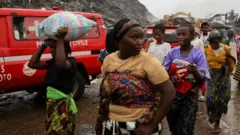 Akitan kan ga gogoro ninu aworan yii, bẹẹ ni awọn obinrin kan n fi ẹṣẹ rin lọ, awọn kan pọn ọmọ sẹyin ninu wọn, bẹẹ ni ẹnikan gbe apo ru sori. Ẹrọ katkata wa lẹyin, bẹẹ ni ọkọ pupa kan naa wa nibẹ pẹlu.