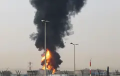 Fumaça e chamas sobem de uma área próxima ao Aeroporto Internacional de Dubai, nos Emirados Árabes Unidos. Alguns prédios baixos podem ser vistos, cercados por uma cerca de segurança com o que parece ser arame farpado
